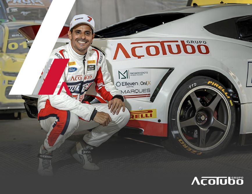 alt="Julio Campos, piloto da Stock Car, com o seu uniforme ao lado do carro branco e vermelho com o logo da parceira Açotubo"