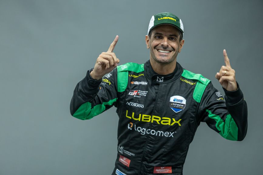 alt="Julio Campos, piloto da Stock Car, com o seu uniforme da Stock Car com os dedos apontando para cima indicando áurea de vitória"